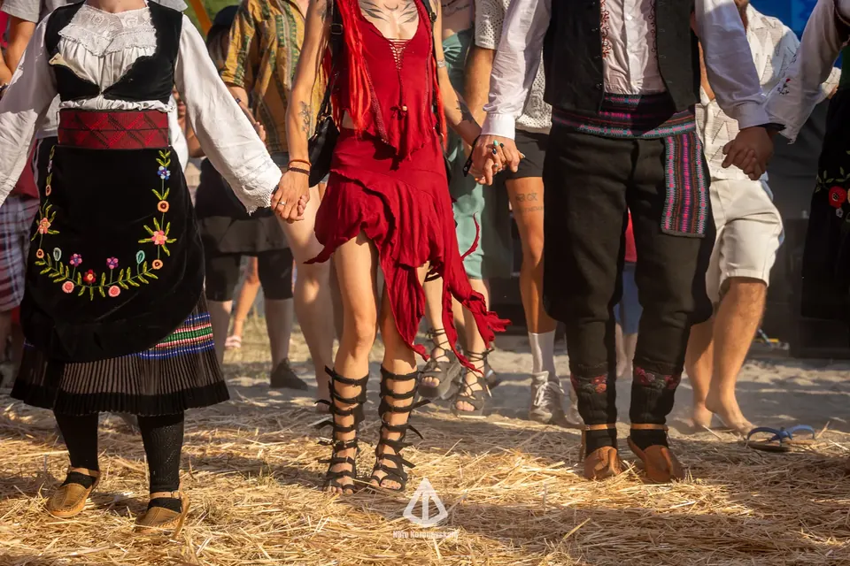 Krupni plan spojenih ruku i stopala u KOLU, tradicionalne vezene nošnje pored festivalske odeće na tlu prekrivenom slamom.