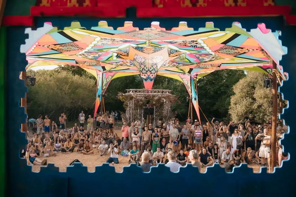 Festival guests gathered under a colourful canopy stage, seen through a carved wooden window