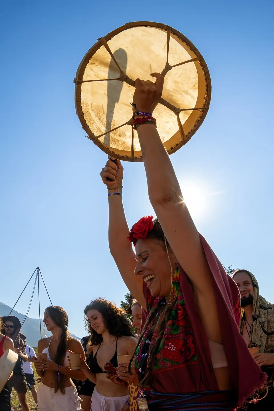A woman raises a frame drum towards the sun
